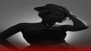 Fotografia artística em preto e branco de uma mulher com expressão séria e pose impactante. Ela usa um turbante estampado na cabeça, brincos longos e um top preto amarrado ao pescoço. Seu rosto está decorado com pinturas decorativas simbólicas, e ela leva a mão à testa, transmitindo uma sensação introspectiva e espiritual. A imagem é iluminada por uma luz suave, destacando suas feições e a atmosfera culturalmente rica, simbolizando o que é quimbanda.