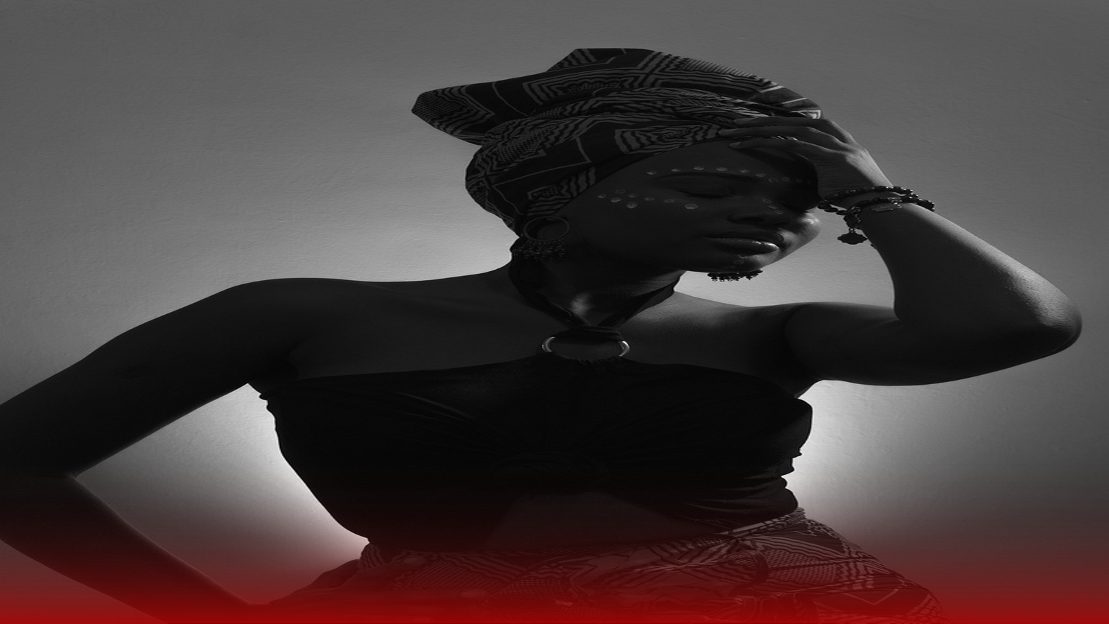 Fotografia artística em preto e branco de uma mulher com expressão séria e pose impactante. Ela usa um turbante estampado na cabeça, brincos longos e um top preto amarrado ao pescoço. Seu rosto está decorado com pinturas decorativas simbólicas, e ela leva a mão à testa, transmitindo uma sensação introspectiva e espiritual. A imagem é iluminada por uma luz suave, destacando suas feições e a atmosfera culturalmente rica, simbolizando o que é quimbanda.