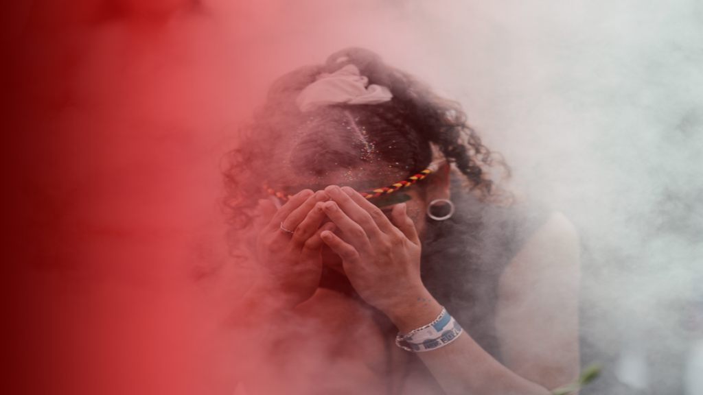 Uma mãe de santo com as mãos cobrindo o rosto, envolta em uma névoa branca e tons de vermelho no fundo. Usa um adereço colorido na cabeça, remetendo a elementos espirituais ou rituais. A imagem transmite mistério e espiritualidade e representa o ritual de como trazer o ex de volta de forma definitiva. 