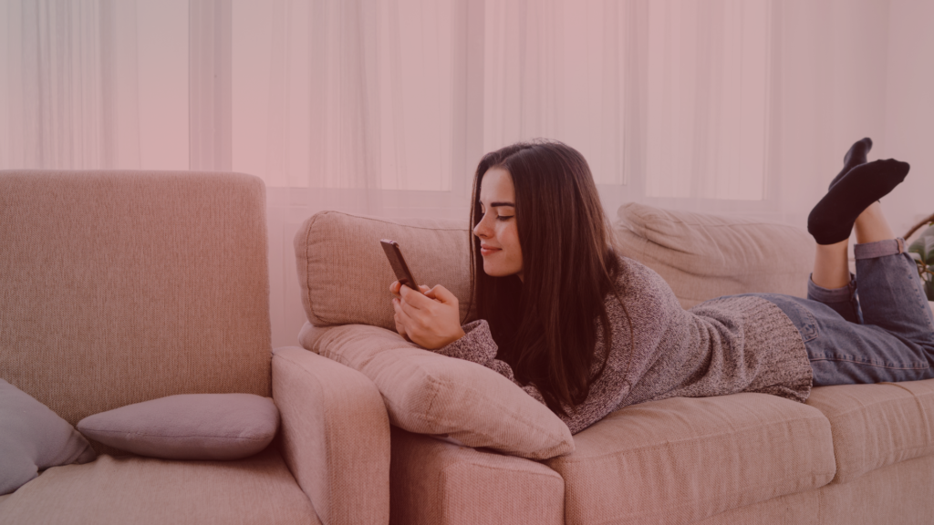 Mulher deitada confortavelmente no sofá, sorrindo enquanto mexe no celular. A cena transmite tranquilidade, mas o contexto sugere possíveis atitudes impulsivas, como enviar diversas mensagens para tentar reconquistar o ex dela.