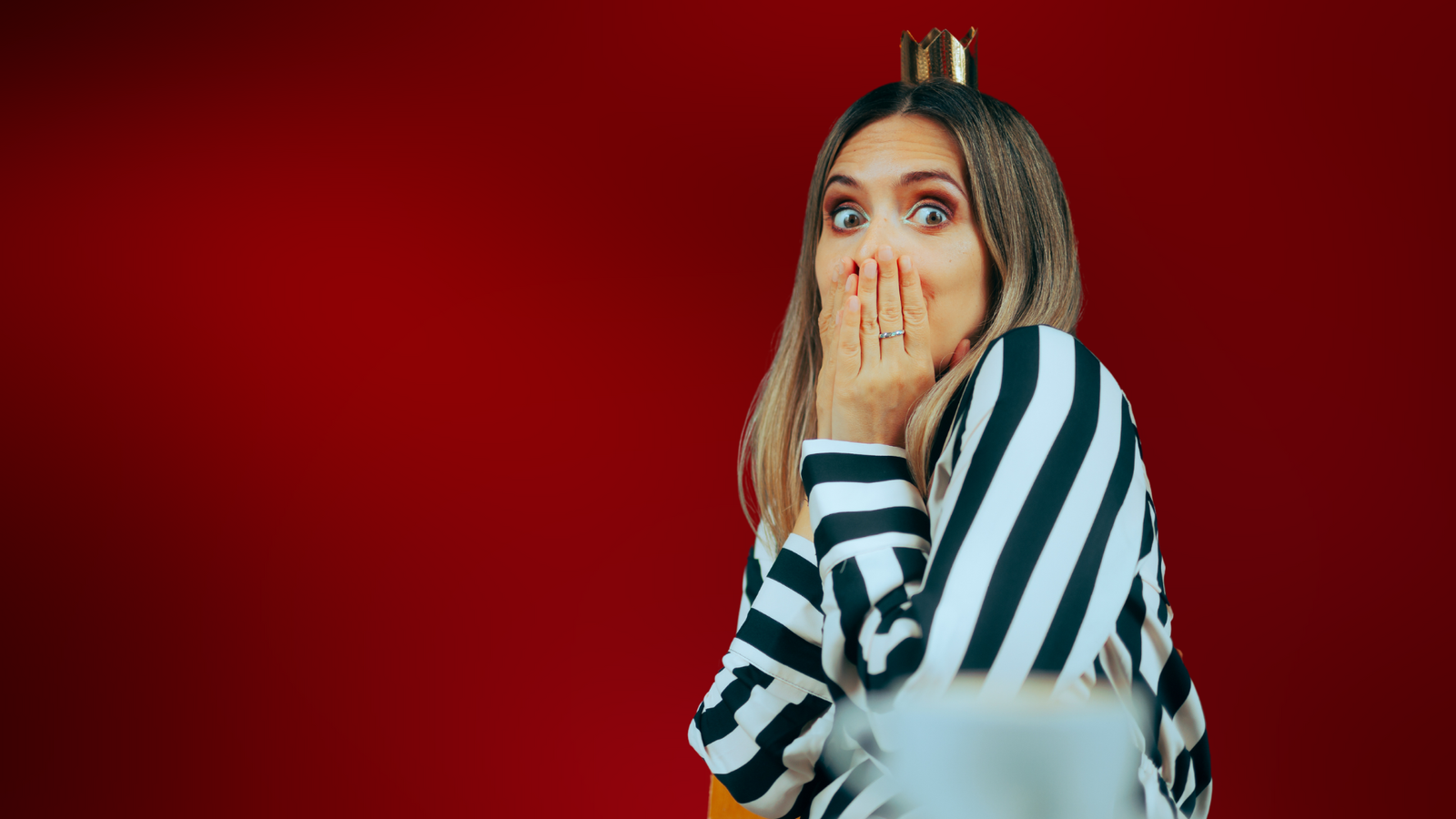 Mulher com expressão surpresa, usando uma coroa dourada em miniatura sobre a cabeça. Ela veste uma blusa listrada em preto e branco e cobre a boca com as mãos, em um fundo vermelho vivo. A imagem transmite a sensação de que a mulher cometeu um erro ao tentar reconquista o ex dela.