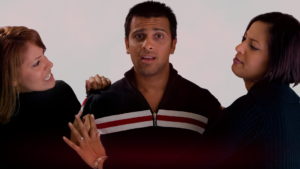 Homem com expressão confusa e preocupado no centro da imagem, sendo segurado por duas mulheres vestidas de preto. A mulher à esquerda parece irritada e está gesticulando de forma agressiva, enquanto a mulher à direita tem uma expressão mais tensa e está segurando o braço do homem. O fundo é neutro, destacando o trio e suas expressões faciais intensas, simbolizando como afastar uma rival.