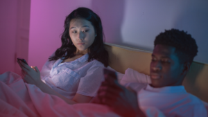 Um casal deitado na cama, ambos usando celulares. A mulher olha para o homem com uma expressão de desconfiança e preocupação, enquanto ele está focado no celular. A iluminação suave em tons de rosa e azul cria uma atmosfera íntima e reflexiva. A cena sugere um possível momento de ciúmes ou desconforto no relacionamento.