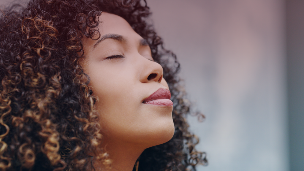 Uma mulher de cabelos cacheados e volumosos aparece em um momento de tranquilidade, com os olhos fechados e uma expressão serena no rosto. A iluminação suave e o fundo desfocado criam uma sensação de paz e autoconfiança. A imagem transmite a ideia de equilíbrio emocional, controle dos sentimentos e bem-estar interior.