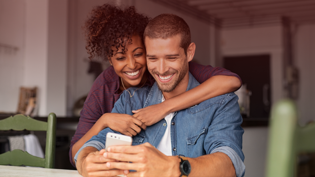 Um casal sorridente compartilha um momento de alegria enquanto olha para o celular. A mulher abraça carinhosamente o parceiro por trás, demonstrando cumplicidade e confiança. O ambiente aconchegante e descontraído reforça a sensação de uma relação saudável e equilibrado, onde há segurança emocional e comunicação aberta.