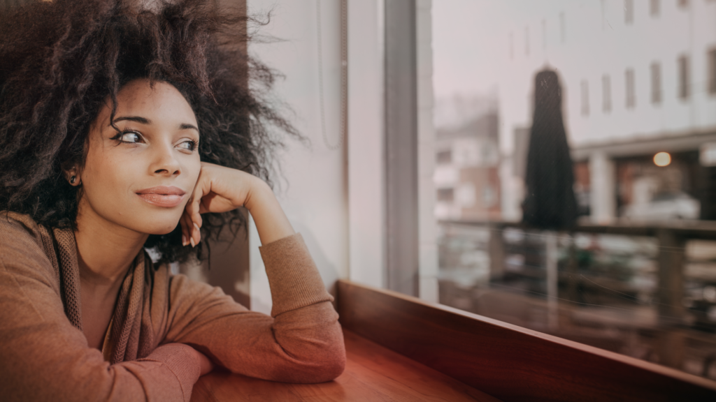 Mulher olhando pensativamente para fora de uma janela, com uma expressão tranquila e reflexiva, em um ambiente interno aconchegante. Simbolizando que ela está a sonhar com ex