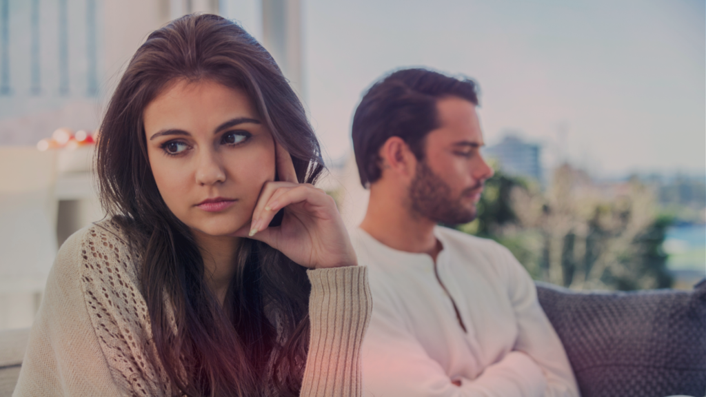 Mulher com expressão pensativa e preocupada em primeiro plano, olhando para o lado, enquanto um homem ao fundo está virado de costas, com os braços cruzados e uma expressão fechada. O ambiente é iluminado e aconchegante, mas o casal parece distante emocionalmente, sugerindo tensão ou desentendimento.