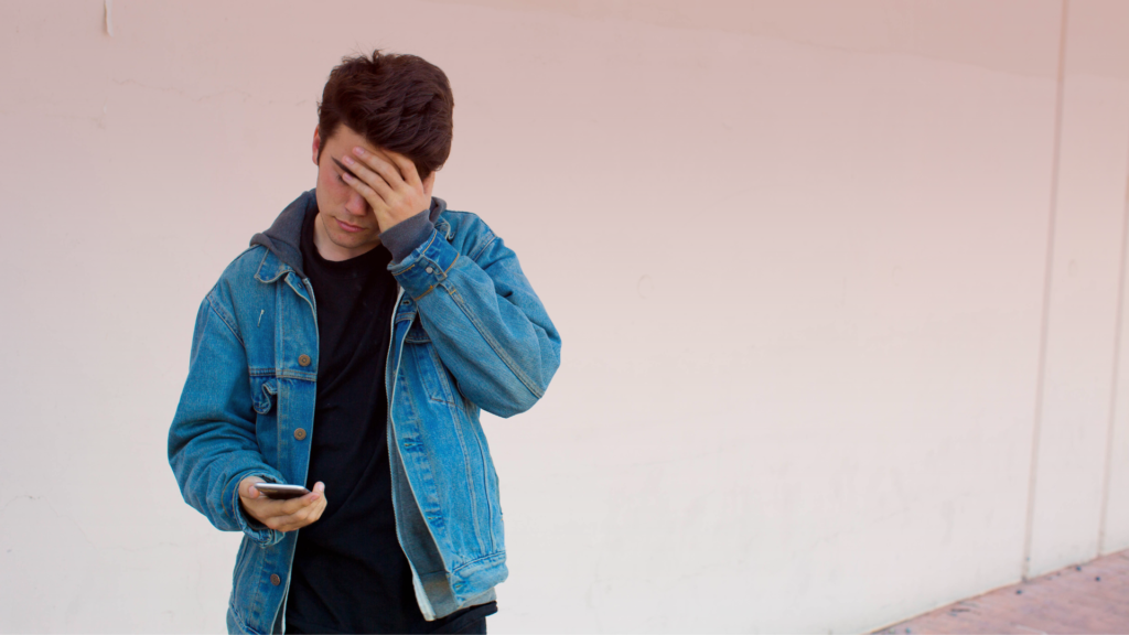 Jovem com expressão preocupada segura um celular enquanto cobre parte do rosto com a mão, transmitindo frustração ou tristeza. O fundo neutro e a postura sugerem um momento de desânimo ou reflexão, simbolizando que ele bloqueou a ex