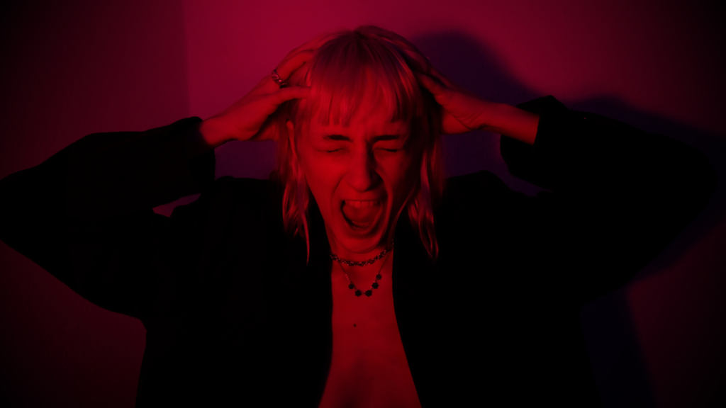 Mulher segurando a cabeça com as mãos e gritando, expressando frustração ou desespero. A iluminação em tons avermelhados e o fundo escuro criam uma atmosfera intensa e emocional, simbolizando o que não fazer quando ele te bloqueou.
