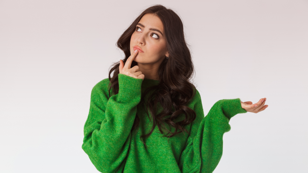 Mulher de suéter verde com expressão pensativa, tocando o queixo com um dedo enquanto olha para cima, sugerindo dúvida ou reflexão. Sua outra mão está levantada em um gesto questionador.