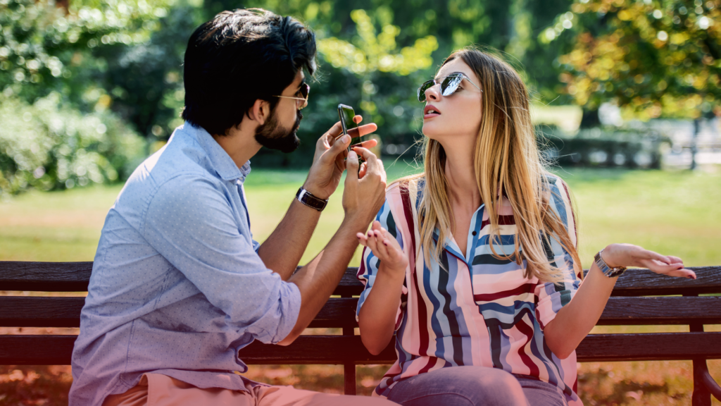 Um casal sentado em um banco de parque durante o dia discute enquanto o homem segura um celular na frente da mulher. Ele parece questionador, enquanto ela, de óculos escuros, gesticula com expressão de impaciência ou defesa. O ambiente ao redor é iluminado e natural, contrastando com a tensão emocional entre eles. A cena sugere um momento de aflição, mas também retrata a dinâmica do relacionamento e o impacto desse sentimento na comunicação do casal.