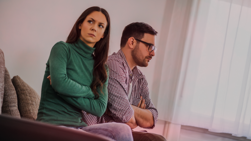 Casal sentado no sofá, ambos os braços cruzados, demonstrando insatisfação e distanciamento emocional. A mulher olha para o lado com uma expressão de frustração, enquanto o homem olha para frente, aparentemente pensativo e desanimado. A cena transmite um relacionamento desgastado pela rotina.