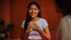 Uma jovem sorridente, com cabelos longos e escuros, usando uma blusa clara, com as mãos sobre o peito em um gesto que expressa emoção e sinceridade, percebendo os sinais de amor. Ela está sentada em um ambiente aconchegante, com uma parede de tom avermelhado ao fundo, conversando com outra pessoa que aparece parcialmente na imagem.