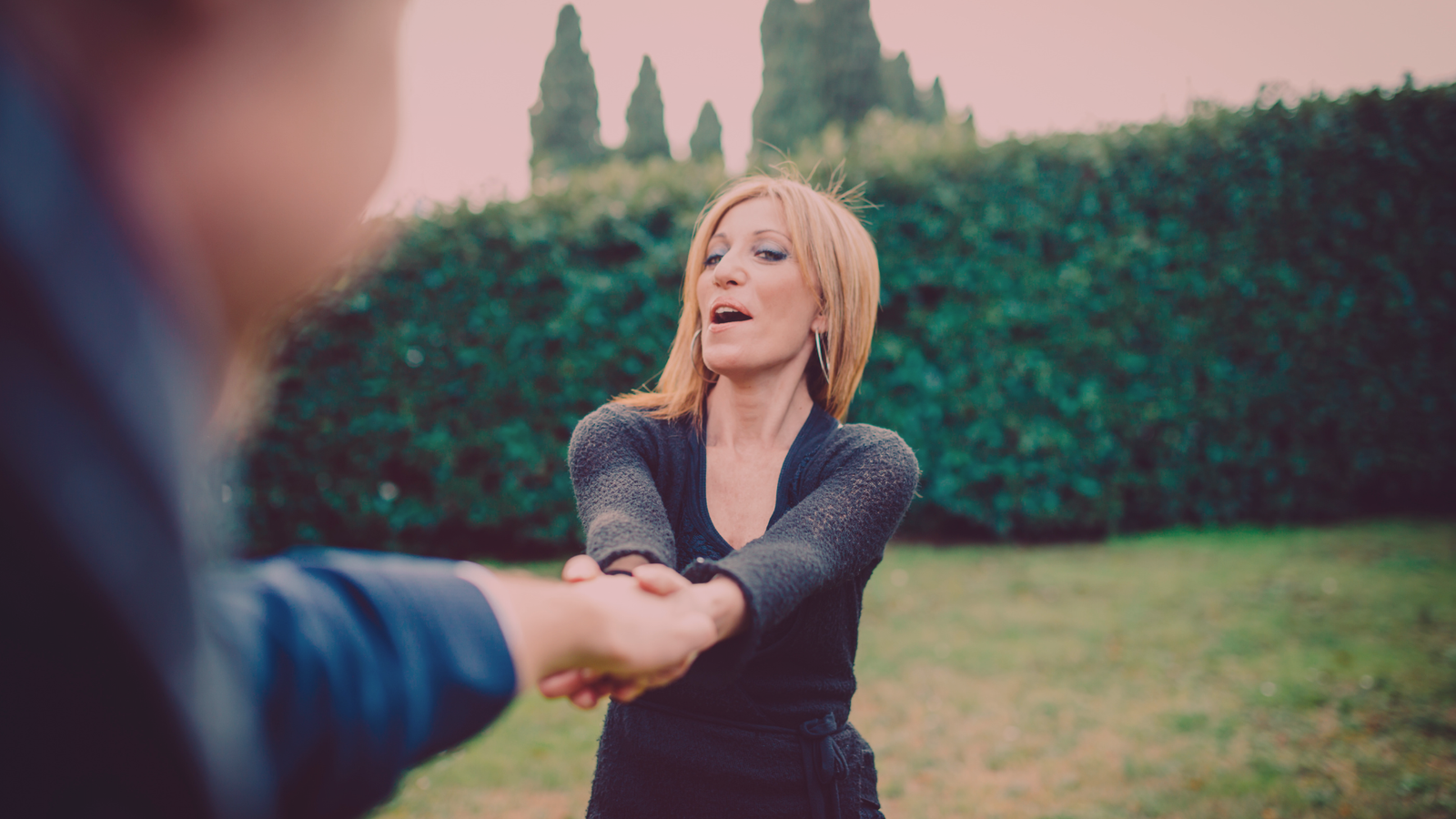 Mulher de expressão confiante segurando as mãos de um homem desfocado no primeiro plano, ao ar livre, com vegetação ao fundo. A cena transmite a ideia de que a mulher está a correr atrás do homem, sugerindo um jogo de conquista e atração.