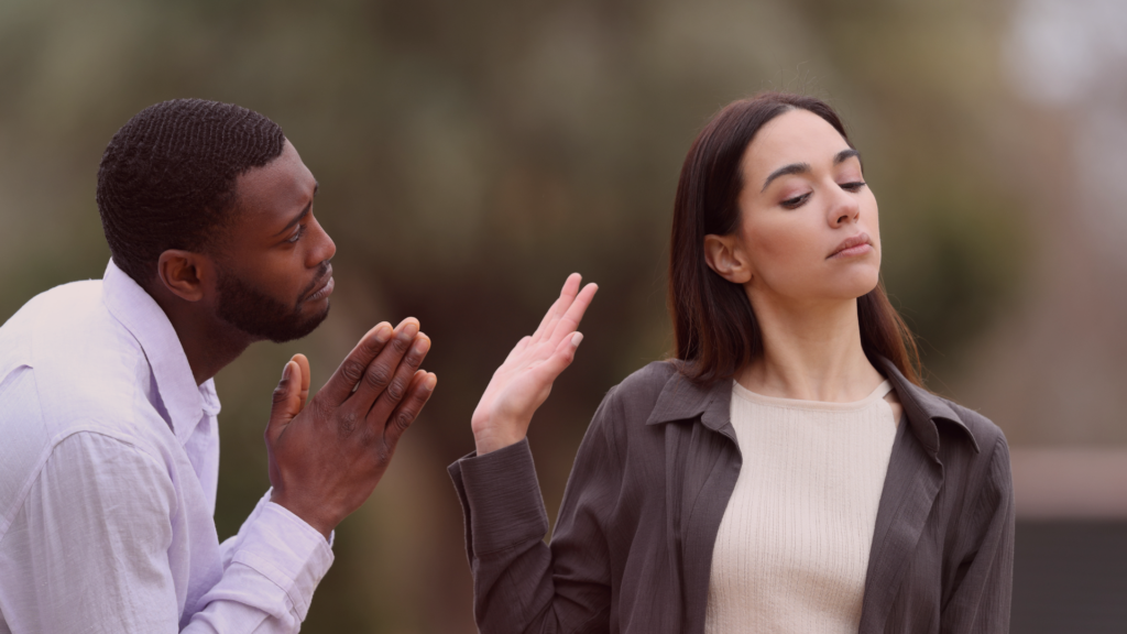 Homem de expressão arrependida juntando as mãos em súplica enquanto olha para uma mulher que mantém um semblante indiferente e levanta a mão em um gesto de recusa. A cena simboliza uma mudança na dinâmica do relacionamento, onde a indiferença desperta o interesse e faz o outro correr atrás.