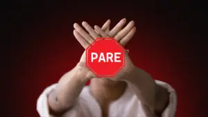 Pessoa com as mãos cruzadas em forma de “X” à frente do rosto, com um sinal de "pare" vermelho sobreposto, simbolizando a decisão firme de contato zero. Fundo em tom escuro com vermelho em destaque, transmitindo urgência e firmeza.