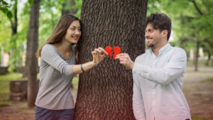 Homem e mulher sorrindo um para o outro enquanto seguram, juntos, as metades de um coração vermelho partido, simbolizando a tentativa de reconciliação em um relacionamento difícil. Eles estão em lados opostos de uma árvore em um parque, o que transmite a ideia de obstáculos, distância e reconexão.