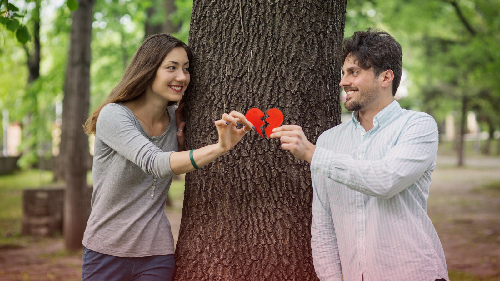 Homem e mulher sorrindo um para o outro enquanto seguram, juntos, as metades de um coração vermelho partido, simbolizando a tentativa de reconciliação em um relacionamento difícil. Eles estão em lados opostos de uma árvore em um parque, o que transmite a ideia de obstáculos, distância e reconexão.
