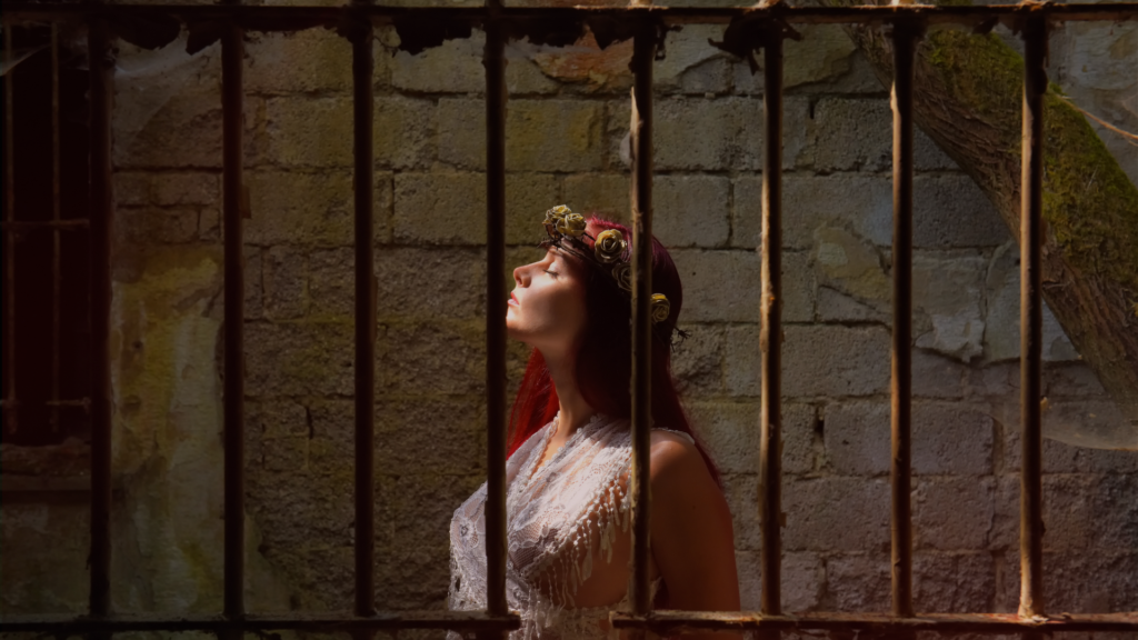 Mulher com expressão serena dentro de uma cela com grades, olhando para cima com os olhos fechados, como se buscasse luz ou esperança — simbolizando o despertar para a liberdade afetiva.