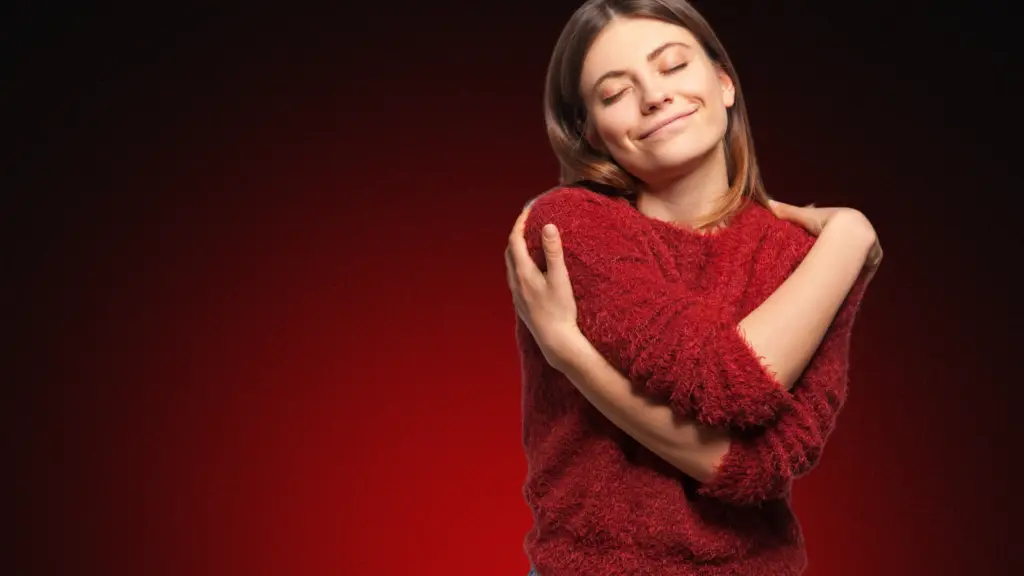 Mulher sorridente com os olhos fechados, abraçando a si mesma com ternura. Ela transmite paz interior, autoconfiança e bem-estar emocional, depois do contato zero com ex. O fundo em tons vermelhos e escuros destaca ainda mais o sentimento de fortalecimento pessoal.
