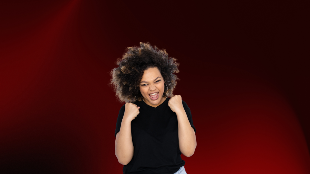Mulher jovem com expressão de alegria intensa e punhos cerrados em sinal de vitória ou empolgação, depois de encontrar a pessoa certa. Ela sorri de forma expansiva, transbordando felicidade e energia. Veste uma camiseta preta e está diante de um fundo vermelho escuro que realça sua presença. 