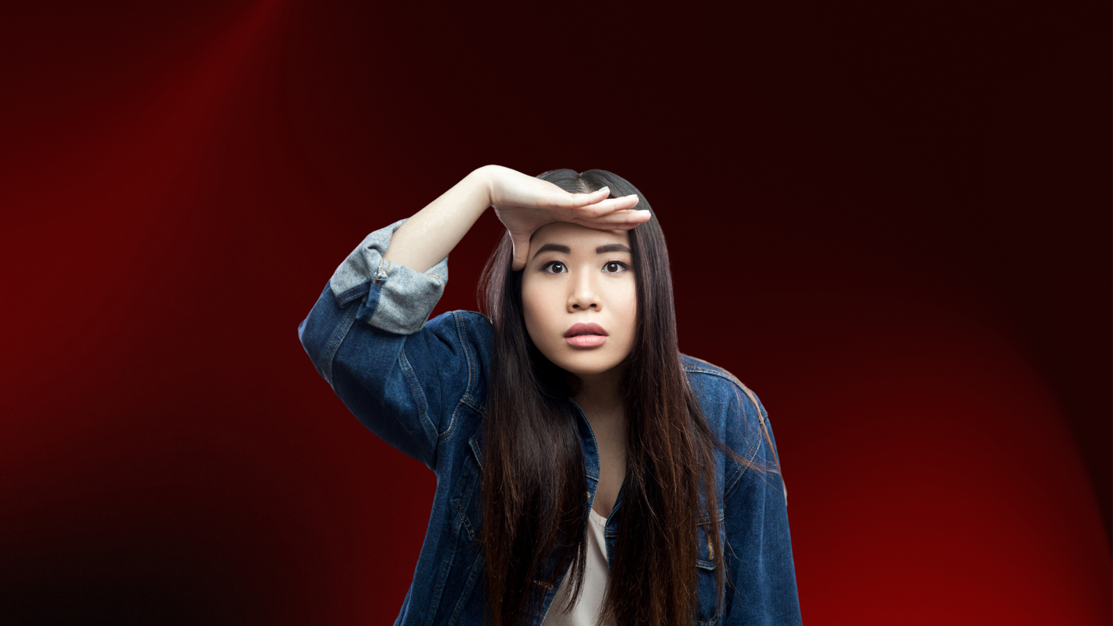 Mulher olhando à distância com expressão de busca, cobrindo os olhos com a mão como se procurasse algo ou alguém. Usa uma jaqueta jeans sobre uma blusa clara, em fundo vermelho escuro. Representa o conceito de "procurar a pessoa certa" com intensidade emocional e expectativa.