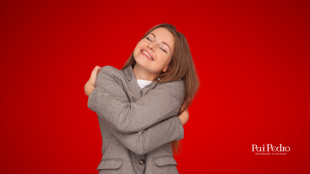 Mulher sorridente de olhos fechados, abraçando a si mesma com ternura diante de um fundo vermelho. A imagem expressa autoestima, acolhimento e a integração da vibração das mulheres.