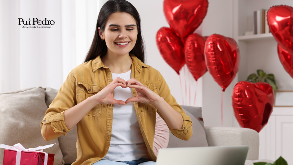 Mulher sorridente faz gesto de coração com as mãos em frente ao notebook, cercada de balões em formato de coração, simbolizando namoro à distância.
